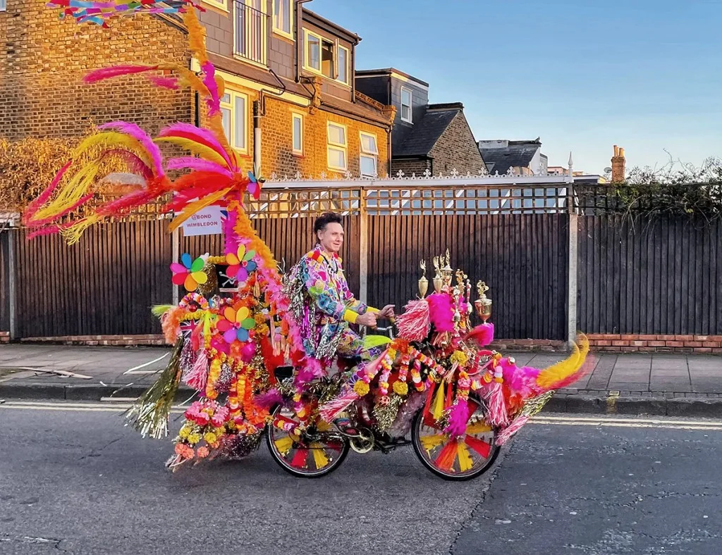 street photo of a man wearing colourful clothes riding a bike with colourful feathers and other objects attached