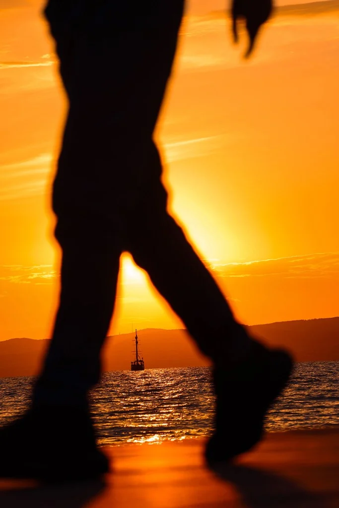 sunset photo looking through a silhouette of someone's legs at a sailing boat