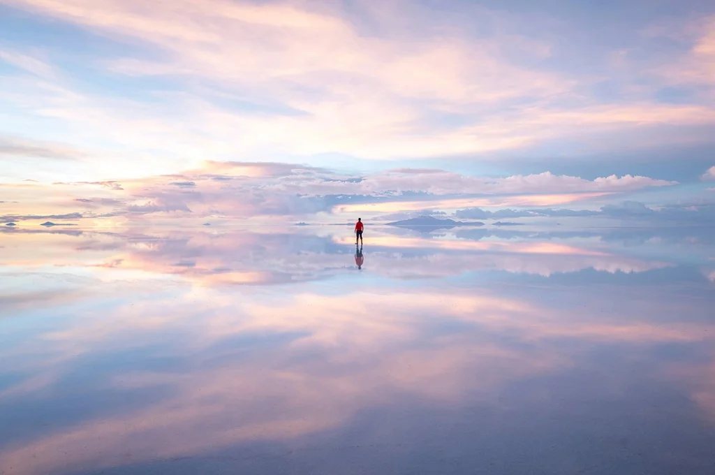 single person standing in lilac and pink sunrise scene reflected in the water