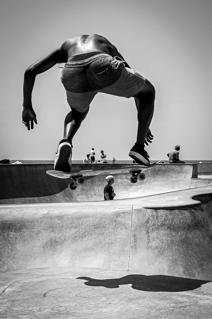 A skateboarder performs a jump infront of the photographer and whilst suspended in midair above the bowl his board is seemingly balanced on another skater’s head below.