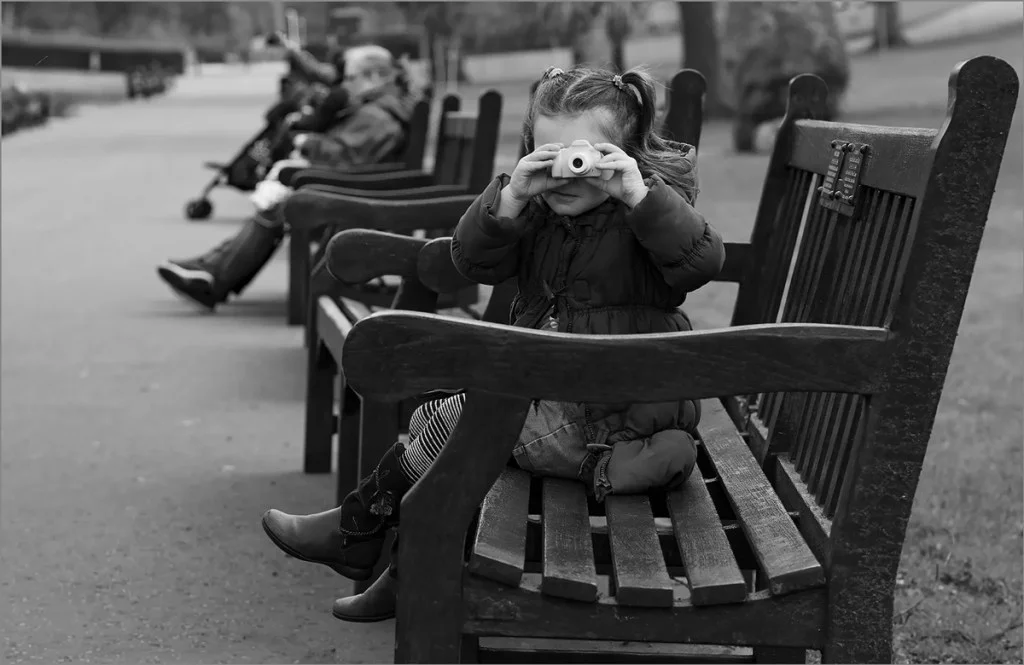 young girl sat on a bench holding up a camera