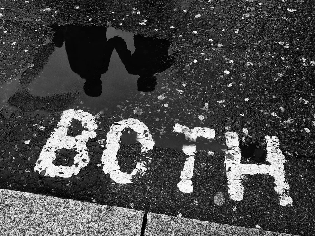 Photo of the word 'both' on the ground next to a puddle with a reflection of two people